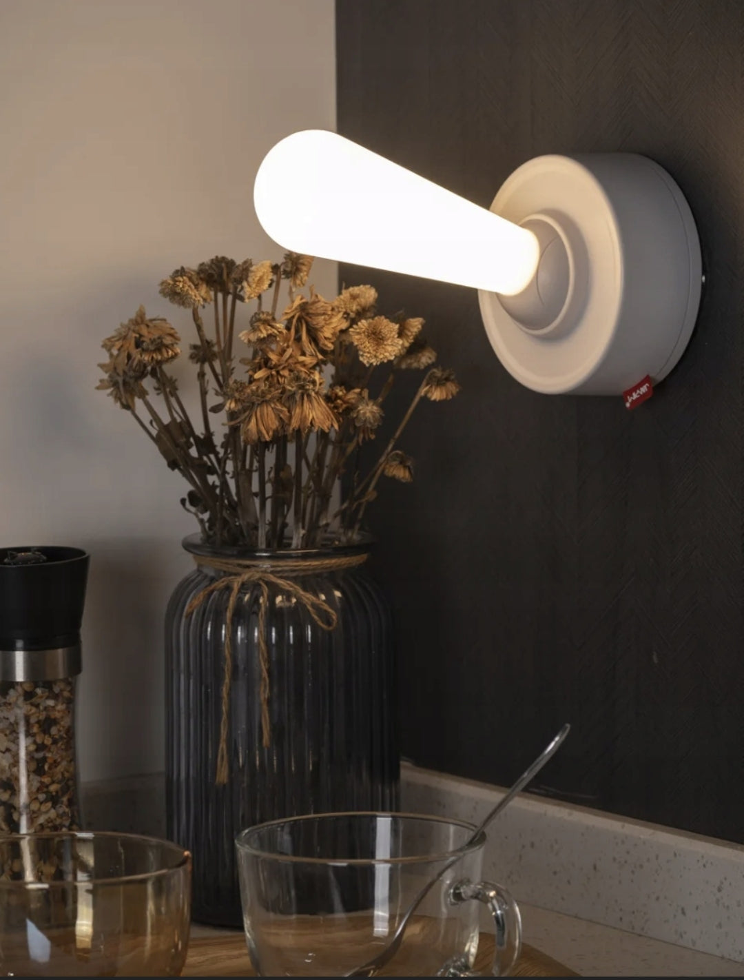 Wall-mounted light fixture on a dark wall with a vase of dried flowers and glasses on a table.